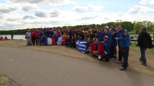 Viele Schüler unterschiedlicher Nationen posieren für ein Gruppenfoto am Werdersee, einige halten Flaggen hoch.