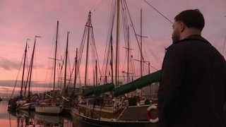 Ein Mann schaut auf Schiffe in einem Hafen während eines Sonnenunterganges. 