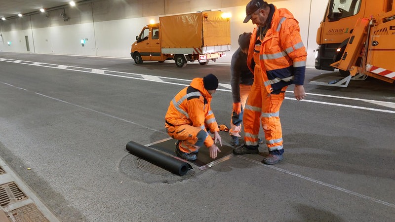 Bauarbeiter stehen vor Baufarzeugen in einem leeren Tunnel und arbeiten am Boden.