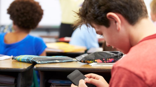 Ein Schüler tippt heimlich während des Unterrichts eine Nachricht auf sein Mobiltelefon.