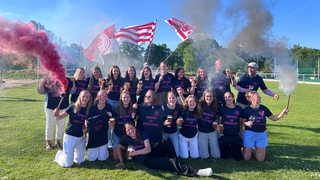 Die Spielerinnen des Bremer Hockey-Clubs posieren ausgelassen beim Mannschaftsfoto auf dem Rasen nach dem Aufstieg und schwenken dabei Vereinsfahren und zünden roten Rauch.