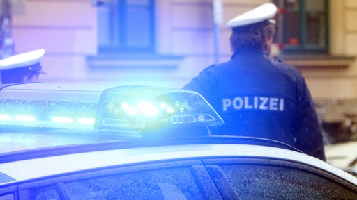 Ein Polizist in Uniform steht vor einem Einsatzwagen der Polizei mit Blaulicht. (Symbolbild)