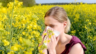 Eine Junge Frau steht in einem gelb blühenden Feld und niest in ein Taschentuch.