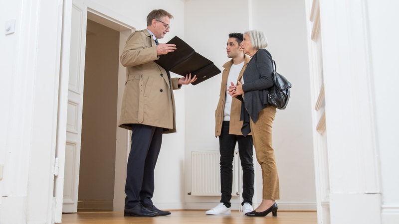 Ein Makler zeigt einem jungen Mann und einer älteren Frau eine leerstehende Wohnung