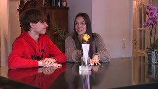 Luna Albanese und Dimitri Kalistov sitzen Zuhause mit dem WM-Pokal der Junioren-WM vor sich.