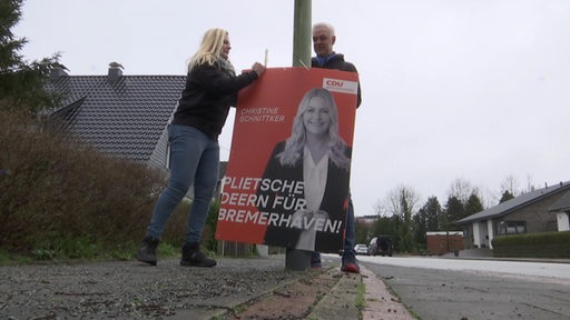 Zwei Wahlhelfer befestigen ein Wahlplakat an einer Laterne.