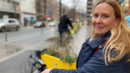 Eine Frau mit langen blonden Haaren steht an der Martinistraße und schaut in die Kamera.