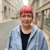 Eine junge Frau mit pink-roten Haaren schaut geradeaus in die Kamera und lächelt leicht.