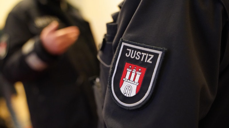 Auf dem Ärmel einer dunklen Uniform ist ein Wappen mit einem Tor und der Aufschrift Justiz abgebildet.