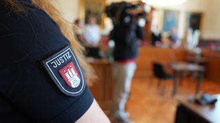 Eine Justizbeamtin steht im Sitzungssaal, auf ihrem Arm steht "Justiz".