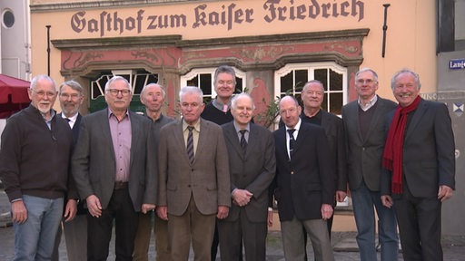 Viele Männer stehen in Anzug vor einem Gasthof.