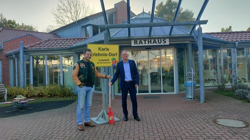 Zwei Männer stehen mit Spaten und Schild vor einem Gebäude.