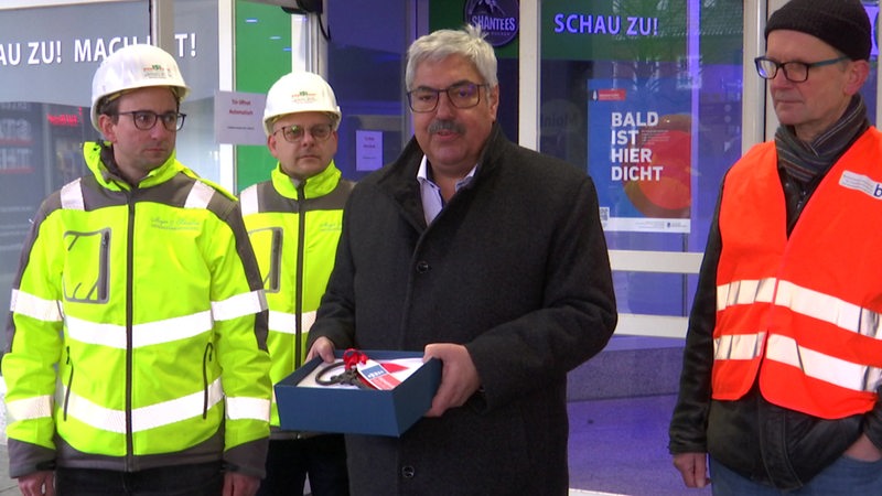 Ein Mann im Mantel steht zwischen Männern mit Warnwesten und hält eine Schachtel in der Hand.