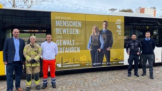 Neben einem Bus mit der Aufschrift "Menschen bewegen ohne Gewalt erfahren" stehen fünf Personen.