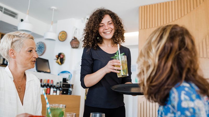 Eine junge Frau serviert einen einen Cocktail in einer Bar. 