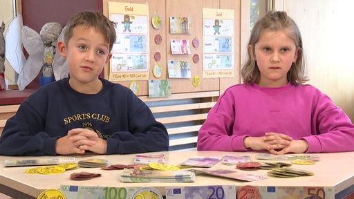 Zwei Kinder sitzen hinter einem Tisch voller Spielgeld