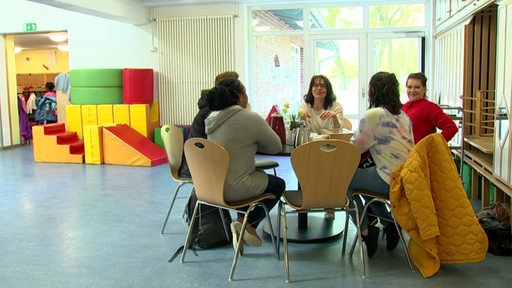 In einem großen hellen Raum sitzen mehrer Personen an einem Rundentisch und unterhalten sich. Im Hintergrund sind bunte Spielsachen zu sehen. 