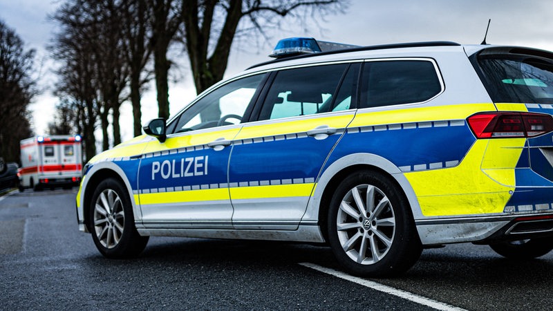  Ein Einsatzfahrzeug, Streifenwagen, der Polizei steht mit Blaulicht und dem Schriftzug als Straßensperre auf einer Landstraße. Im Hintergrund sieht man einen Rettungswagen, RTW an einer Unfallstelle stehen. 