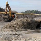 Vor einem beschädigten Deich schießt ein Wasser-Sand-Gemisch aus einem Rohr, dahinter steht ein Bagger bereit. 