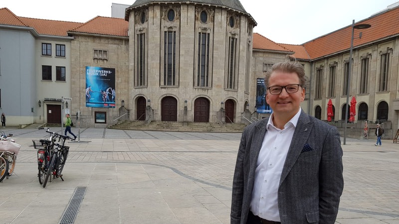 Ein Mann im Anzug steht vor einem Thetaer Gebäude.