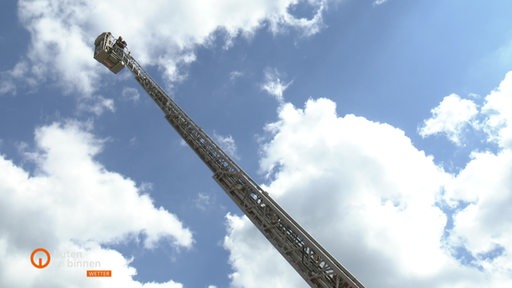 Zwei Menschen sind auf einer Drehleiter der Bremer Feuerwehr zu sehen. Über ihnen ist der Himme zu sehen.