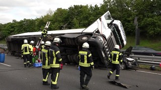 Auf einer Autobahn ist ein Lkw auf einen Pkw gekippt. Im Vordergrund stehen Feuerwehrleute.