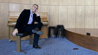 Generalmusikdirektor Marco Comin sitzt lachend auf einem Stuhl vor einem Klavier