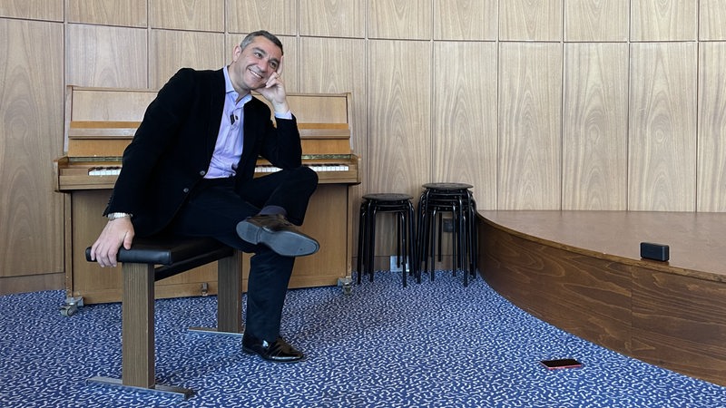 Generalmusikdirektor Marco Comin sitzt lachend auf einem Stuhl vor einem Klavier