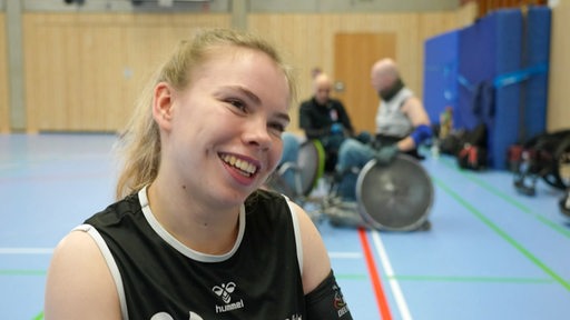 Mascha Mosel wird interviewt. Im Hintergrund sieht man Personen im Rollstuhl in einer Sporthalle sitzen.