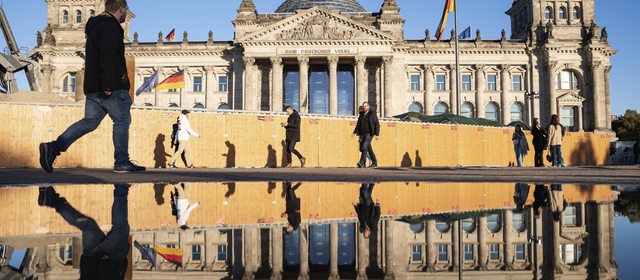 Menschen gehen an einem sonnigen Herbsttag am Reichstag im Berliner Bezirk Mitte vorbei, dessen Westfassade sich in einer großen Pfütze davor spiegelt. 