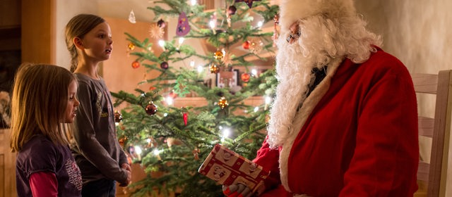 Der Weihnachtsmann sitzt zur Bescherung in einem Haus neben einem geschmückten Weihnachtsbaum während zwei Mädchen ihm ein Lied vorsingen