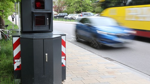 Ein mobiler Blitzer-Anhänger steht an einer Straße, ein Auto und Bus fahren nebenher