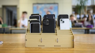 Mehrere Smartphones stehen in einer Holzbox in einem Klassenraum.