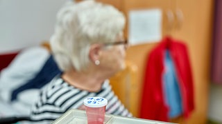 Eine Frau sitzt in einem Zimmer eines Pflegeheims. Im Vordergrund ist eine Medikamentendose zu sehen.