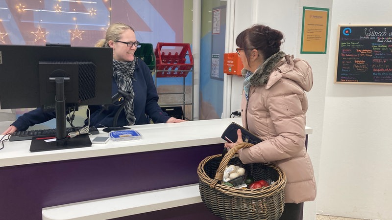 Eine Frau hinter einem Bildschirm spricht mit einer Frau mit einem Einkaufskorb.