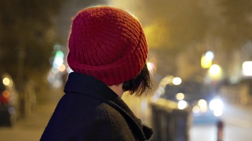 Rückansicht einer Frau mit Mütze, die nachts auf die Straße blickt.