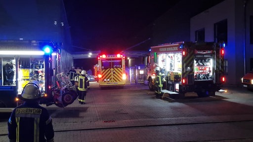 Feuerwehrautos stehen auf einer Einfahrt eines Betriebs. Im Vordergrund steht ein Feuerwehrmann.