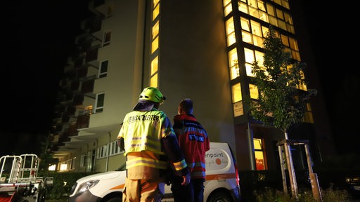 Ein Feuerwehrmann und ein Sanitäter stehen vor einem Hochhaus, in dem ein Feuer ausgebrochen ist.