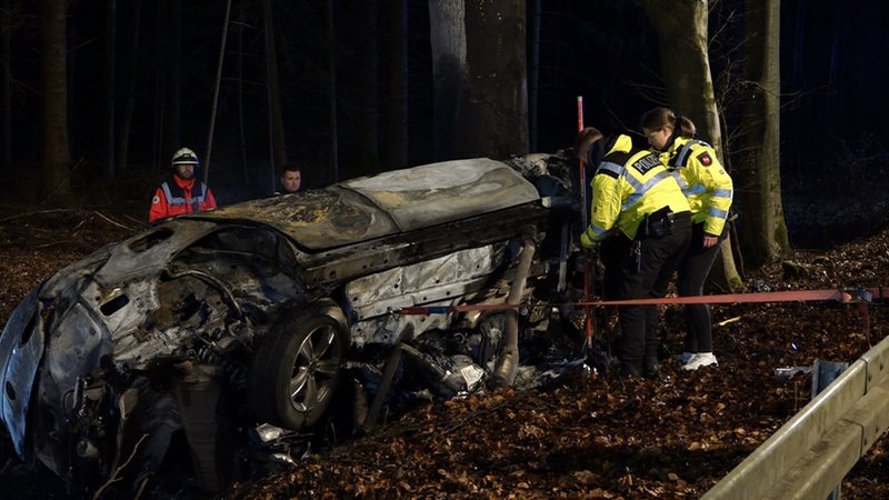 Aus ausgebranntes Auto liegt auf der Seite neben einer Landstraße. Polizisten und Feuerwehrleute stehen daneben.