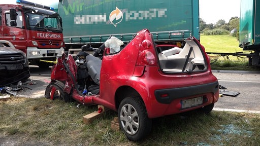 Ein kaputtes Auto steht vor einem Lkw und Feuerwehrauto.