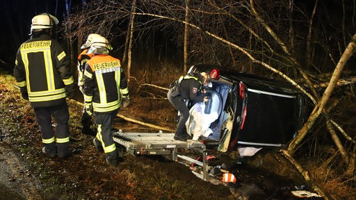 Ein Auto liegt in einem Graben. Eine Polizistin schaut hinein. Daneben stehen Feuerwehrleute.