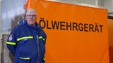 Ein Mann in blauer Uniform steht vor einer orangen Plane mit der Aufschrift "Ölwehrgerät".