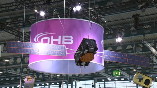 Ein Satellit hängt in einer Halle und im Hintergrund sieht man das OHB Logo.