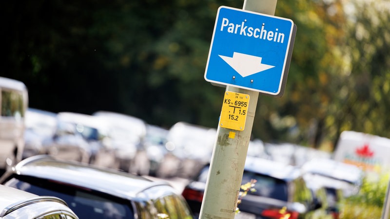 Ein Schild weist am Straßenrand neben geparkten Fahrzeugen auf die Position eines Parkscheinautomaten hin. 