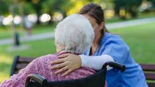 Eine Altenpflegerin hält die Hand auf den Rücken einer Seniorin beim Spaziergang mit Rollator