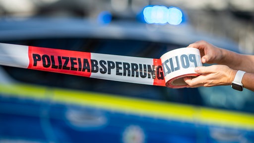 Zwei Hände halten ein rot-weißes-Absperrbabnd mit der Aufschrift Polizeiabsperrung. Im Hintergrund ein Polizeiauto.