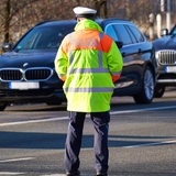 Ein Mann in Polizeiuniform steht mit dem Rücken zur Kamera auf einer Straße. Im Hintergrund sind zwei schwarze BMWs zu sehen.