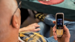 Ein älterer Menschen sitzt in seiner Wohnung und hält ein Mobiltelefon in der Hand. Auf dem Display des Telefons ist die 110 zu lesen.