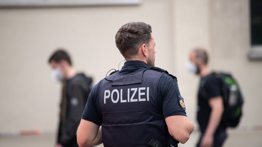 Auf dem Rücken eines Mannes steht "Polizei", der Polizist schaut zur Seite. Im Hintergrund gehen Menschen vorbei.