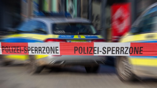 Ein rot-weißes Absperrband ist zu sehen. Darauf steht "Polizei-Sperrzone". Im Hintergrund sind Polizeiautos zu sehen.